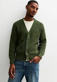 Cardigan verde lavorato a maglia con scollo a V, caratterizzato da un motivo testurizzato e sei grandi bottoni, abbinato a una camicia bianca e jeans blu.