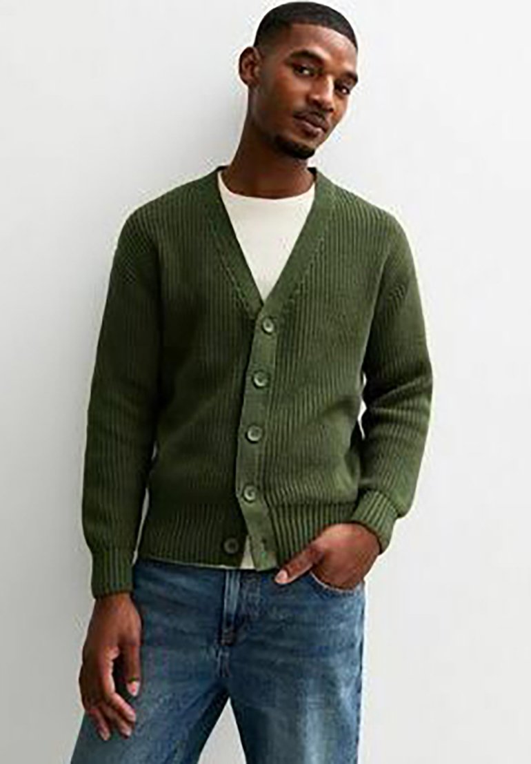 Cardigan verde lavorato a maglia con scollo a V, caratterizzato da un motivo testurizzato e sei grandi bottoni, abbinato a una camicia bianca e jeans blu.