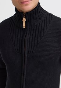 Stickad svart zip-up tröja med hög ribbad krage och en slät dragkedjehandtag med läderdetalj. Strukturerad tyg med en åtsittande design.