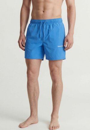 Mann trägt blaue Badehose mit elastischem Bund, steht barfuß auf einfarbigem Hintergrund, die Hose hat kleinen weißen Text am linken Oberschenkel.