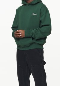 Felpa verde scuro con tasca frontale e logo distintivo ricamato sul petto. Presenta polsini e orlo a costine, indossata con pantaloni cargo neri.