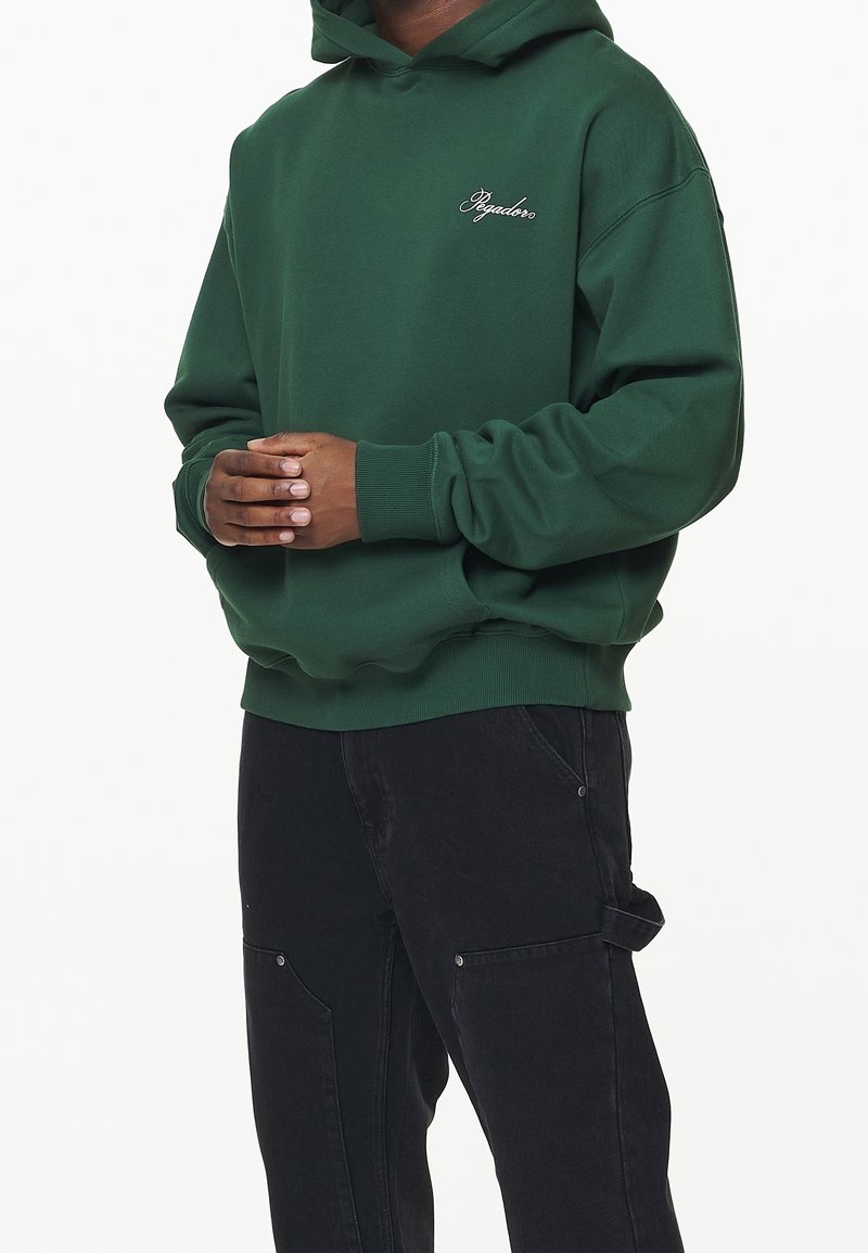 Felpa verde scuro con tasca frontale e logo distintivo ricamato sul petto. Presenta polsini e orlo a costine, indossata con pantaloni cargo neri.