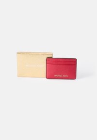 Red leather card holder with textured finish, featuring four card slots. Comes with a metallic gold packaging box with "MICHAEL KORS" label.
