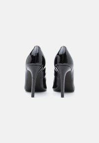 O pereche de pantofi negri din piele lacuită cu toc înalt, prezentați din spate pe un fundal alb.