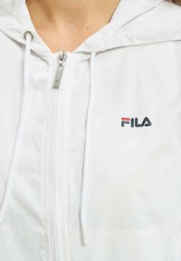 Valkoinen vetoketjullinen takki hupulla, jossa on hopeinen vetoketjuvedin. FILA-logo on mustana ja punaisena edessä. Silkkinen rakenne.