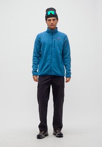 Νέος άνδρας που φοράει μπλε φλις μπουφάν, μαύρο παντελόνι cargo, παπούτσια πεζοπορίας, μαύρο σκουφάκι με ανακλαστικά γυαλιά σκι στην κορυφή, στέκεται μπροστά σε λευκό φόντο.