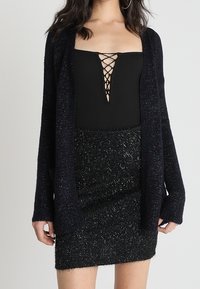 Námořnický dlouhý kardigan s texturovaným povrchem, nošený přes černý top s šněrováním vpředu, kombinovaný s černou třpytivou minisukní.