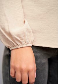 Maglione beige a maniche lunghe con polsini elastici arricciati. Tessuto morbido e strutturato. Mano appoggiata su un tessuto scuro e testurizzato dei pantaloni.