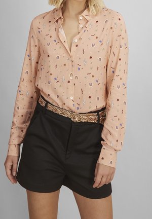 Mujer con camisa color melocotón abotonada con patrones abstractos y coloridos, pantalones cortos negros de talle alto y cinturón trenzado beige con hebilla dorada.
