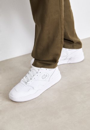 Person iført olivengrønne bukser og hvide atletiksneakers, der står på et beige gulv.