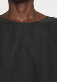 Haut en tissu noir avec une texture lisse et mate et un décolleté large. Le vêtement présente une coupe ample avec des drapés subtils.