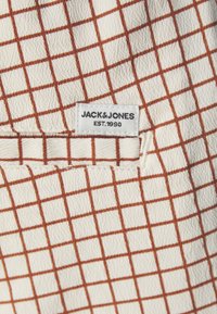 Tejido texturizado con una base color crema que presenta un patrón de rejilla marrón. Incluye una etiqueta blanca con "JACK&JONES EST. 1990" cosida.