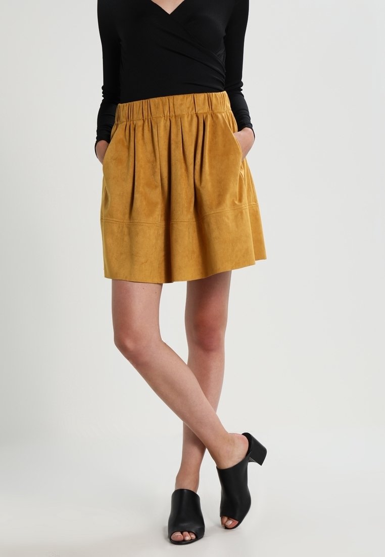 Femme portant une jupe plissée jaune moutarde, un haut noir à manches longues et des mules noires ouvertes à talons, debout les mains dans les poches.
