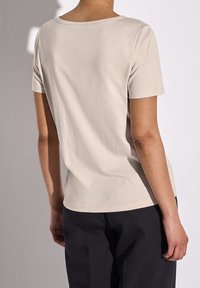Camiseta de manga corta en color beige claro con escote redondeado, de suave tela de algodón y un corte holgado, combinada con pantalones oscuros, vista desde la espalda.