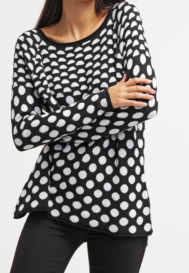 Femme portant un pull noir à manches longues avec de gros et petits pois blancs, associé à un pantalon noir, bras croisés.
