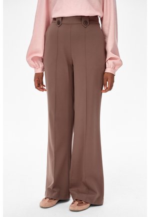 Brązowe spodnie o szerokich nogawkach z wysokim stanem, z przednimi szwami i zapinanymi na guziki klapami, noszone z różową bluzką i pasującymi płaskimi butami.