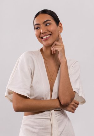 Witte linnenoutfit met een diepe V-hals en korte mouwen, voorzien van een gouden hangerketting. Het model heeft lang haar dat naar achteren is getrokken en glimlacht.