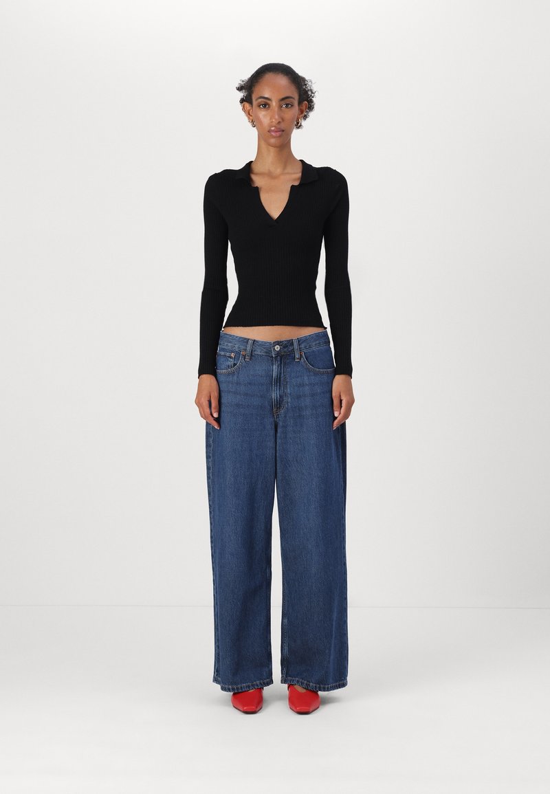 Haut en maille côtelée noire avec col, associé à un jean large en denim bleu. Le mannequin porte des chaussures à talons carrés rouges.