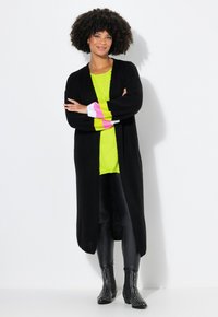 Langer schwarzer Cardigan über einem knallgelben Oberteil, kombiniert mit schwarzen Leggings und besetzten Ankle Boots. Gestreifte, farbenfrohe Akzente an den Ärmeln.