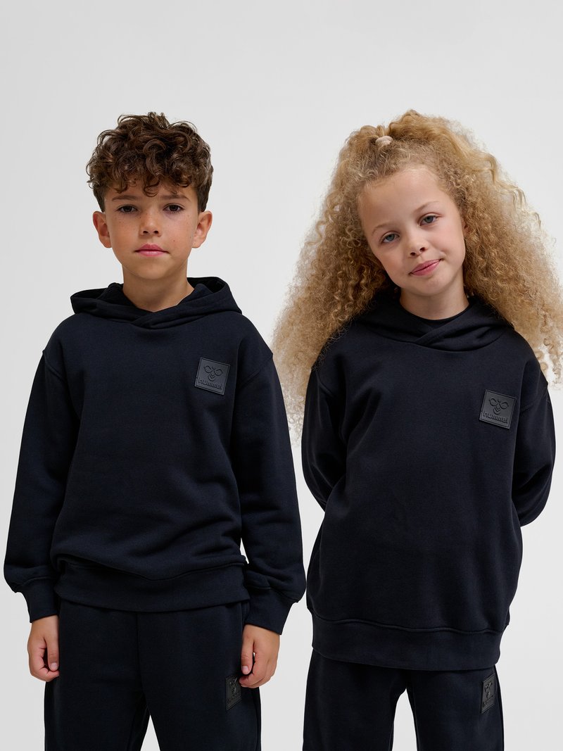Dwoje dzieci stoi obok siebie, ubrane w pasujące czarne bluzy z kapturem i spodnie na jednolitym jasnym tle.