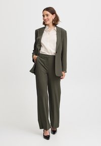 Olivgrüner maßgeschneiderter Anzug mit einreihigem Jacket und geraden Hosen, kombiniert mit einem hellbeigen Hemd mit Knöpfen und schwarzen High Heels.