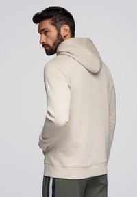 Světle béžová mikina s kapucí na stažení a žebrovanými manžetami. Má jemnou texturu a uvolněný střih, kombinovaná se zelenými kalhotami.