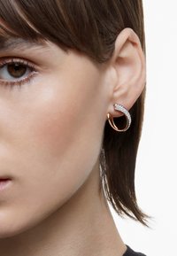 Náušnice vo forme zlatohnedého krúžku s krivkovým dizajnom, zdobené malými trblietavými kryštálmi, na neutrálnom pozadí.