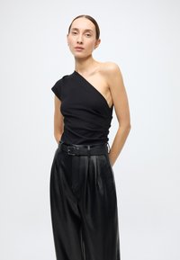 Schwarzes Ein-Schulter-Oberteil aus weichem Material mit drapierten Details, kombiniert mit hoch taillierten schwarzen Lederhosen und einem schlanken Gürtel.