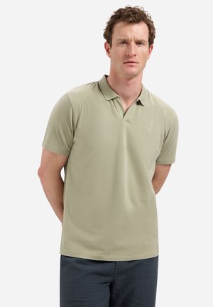 Man met krullend haar die een lichtgroen poloshirt met korte mouwen en donkere, gestructureerde broek draagt, staand met handen achter de rug tegen een witte achtergrond.