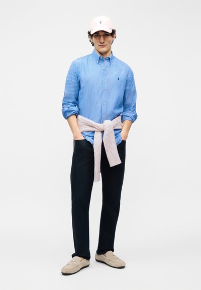 Άνδρας φοράει ανοιχτό ροζ καπέλο, μπλε ριγέ πουκάμισο με κουμπιά, ανοιχτό ροζ πουλόβερ δεμένο στη μέση, μαύρο παντελόνι και μπεζ παπούτσια τύπου slip-on.