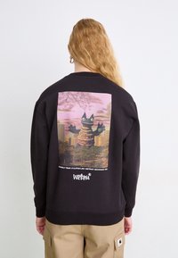 Personne aux longs cheveux blonds porte un sweat-shirt noir arborant un monstre de dessin animé dans un paysage urbain avec un ciel rose dans le dos.