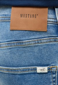 Világoskék farmernadrág barna bőrcímmel, rajta a "MUSTANG" felirattal, a hátsó zseb felett pedig egy fehér szövettag látható. Megerősített varrások láthatók.