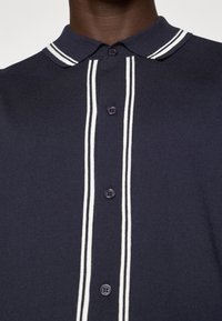 Polo azul-marinho com riscas brancas duplas no colarinho e ao centro. Apresenta uma pala com botões e tecido de malha texturizado. Os botões são de cor roxa escura.