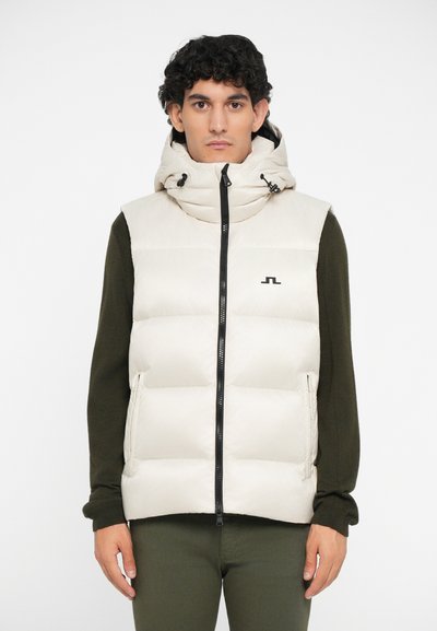 Beige puffer-vest med høj krave og hætte, med diagonalt quiltet design, lynlås og sort logo-detalje. Båret over en mørkegrøn sweater.