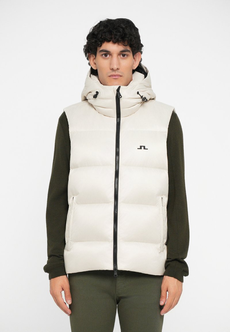 Beige puffer vest with a high collar and hood, featuring a diagonal quilted design, zip closure, and black logo detail. Worn over a dark green sweater.