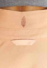 Gros plan sur la zone arrière de la taille d’un pantalon couleur pêche avec un petit motif de feuille brodée et une boucle en tissu au-dessus de la poche.