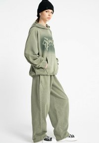 Młoda osoba ubrana w przerośnięty, wyblakły zielony hoodie i dopasowane szerokie spodnie, z czarną czapką beanie i czarno-białymi sneakersami, stojąca bokiem.