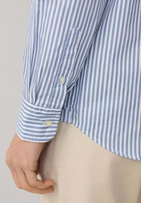 Camisa de rayas azul y blanca con mangas largas, puños abotonados y un dobladillo recto, hecha de una tela ligera. Detalles de botones en los puños.