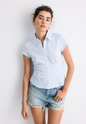 Mladá žena v krátkom rukáve svetlomodrej kockovanej blúzke a opraných rifľových šortkách stojí pred bielym pozadím.