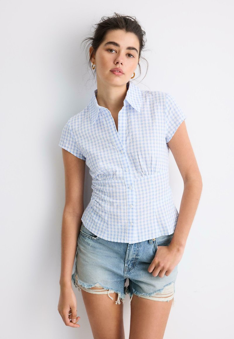 Jeune femme portant un chemisier à manches courtes à carreaux bleu clair et un short en jean usé, debout devant un fond blanc.