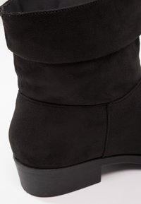 Bottine noire en daim avec un revers plié, surface texturée et un petit talon empilé. Le design est simple avec des lignes épurées et sans embellissements.