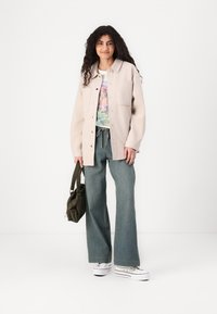 Giacca button-up color beige chiaro con due tasche frontali, maglietta bianca con grafiche, jeans blu larghi in denim e sneakers bianche con plateau. Borsa verde oliva.