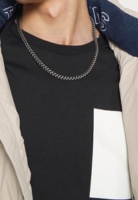 Närbild på en person som bär ett silverhalsband, svart T-shirt med en vit rektangulär design och en beige jacka med en marinblå huva.