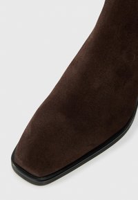 Tmavě hnědá semišová kotníková bota se čtveratou špičkou a černou podrážkou, zobrazená zblízka na bílém pozadí.