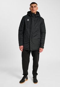 Svart vadderad jacka med huva, försedd med blixtlås och knappstängning, framfickor och en logotyp på bröstet; bärs med svarta byxor och skor.