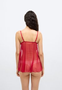 Vrouw met lang donker haar, gekleed in een doorschijnend rood lingerie topje en bijpassende slips, gezien van achteren tegen een eenvoudige lichte achtergrond.