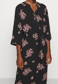 Musta mekko pitkilla hihoilla ja V-p��nteella, jossa on vaaleanpunaisia kukkakuosisia yksityiskohtia. Mekossa on vyötärösolmu, joka luo istuvan ilmeen, sekä nappiyksityiskohtia.