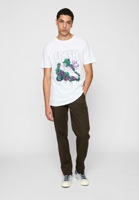 Hvit t-skjorte med et grafisk design av en sko og druer, kombinert med olivengrønne bukser og blåtonede sneakers.