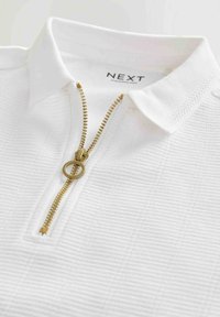 Polo blanc avec un design texturé, une fermeture éclair dorée avec un tirette circulaire et un col pour une allure classique. L'étiquette indique "NEXT."