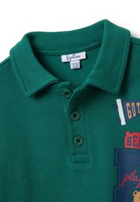 Polo verde in cotone con finitura testurizzata, dotato di colletto, tre bottoni verdi e dettagli in lettering stampati colorati.